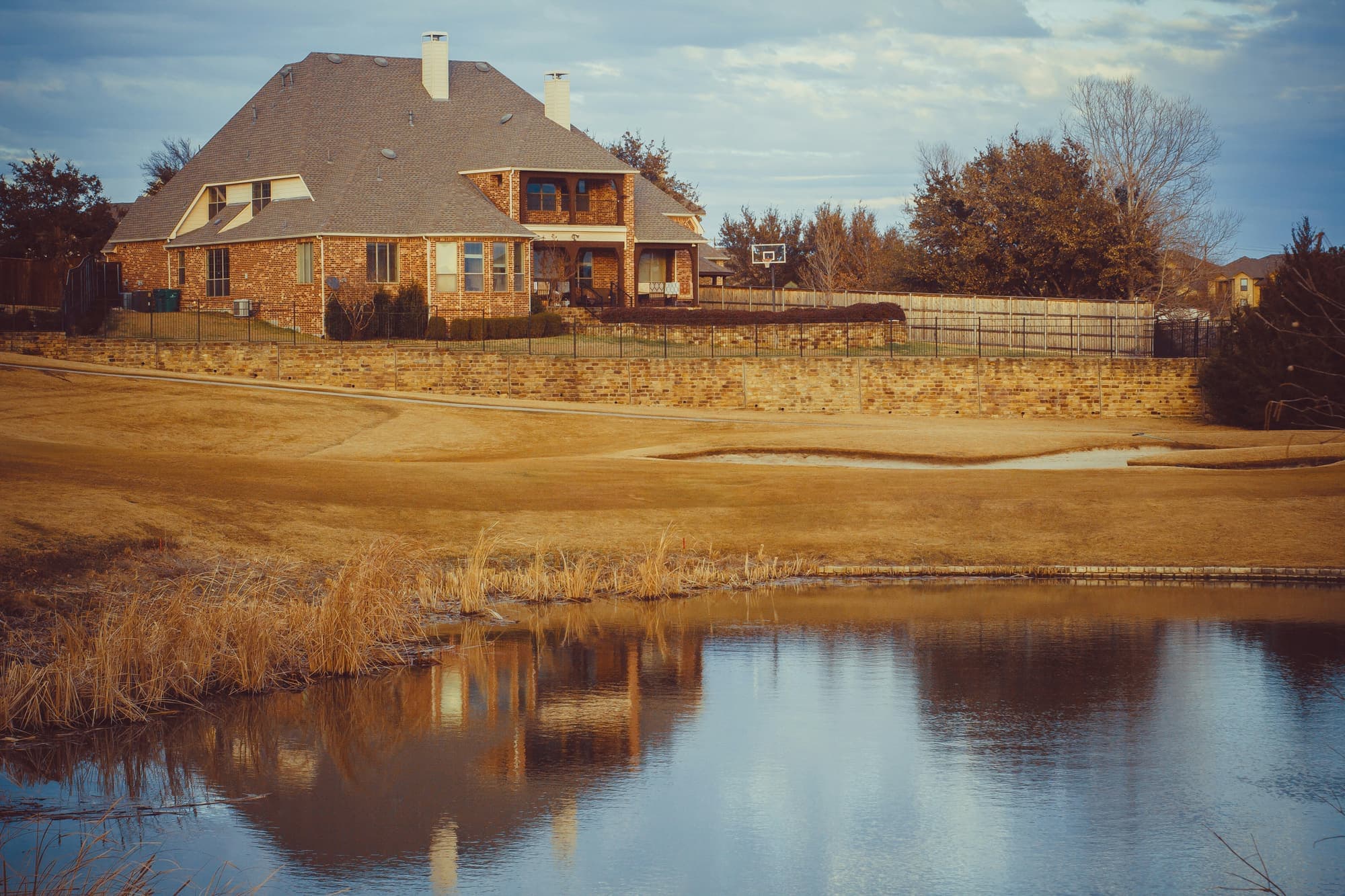 A brick two-story home beside a pond, in warm evening light